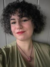 light olive-skinned woman with chin-length curly hair, dark eyes, and red lipstick. She is wearing a light green dress and a pomegranate necklace