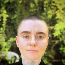 Person with a buzz cut, one nose piercing on each nostril, dimple piercings, a neck tattoo of a magnolia in the center of their throat, standing in front of green foliage. 