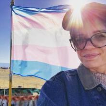 Erika stands in front of a trans flag and an afternoon sun. 