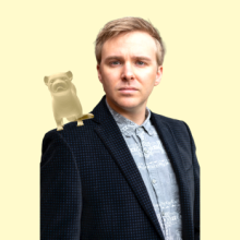 White male with blonde hair, wearing blazer, with 3D ferret animation perched on shoulder.