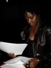 Dark-brown skinned woman looks down at papers in her lap. She is wearing a black top and a leather jacket