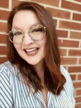 Hannah V Warren, wearing glasses and a striped shirt, has mid-length red hair and is smiling in front of a brick wall.. 