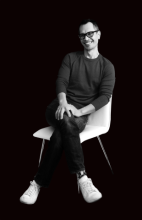 Asian man with black glasses sits on a white chair in front of a black background