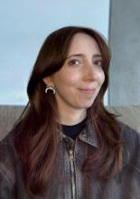 A femme-presenting person with red hair, brown eyes, and white skin sits on a couch wearing a brown jacket and traditional Armenian bullhorn earrings.